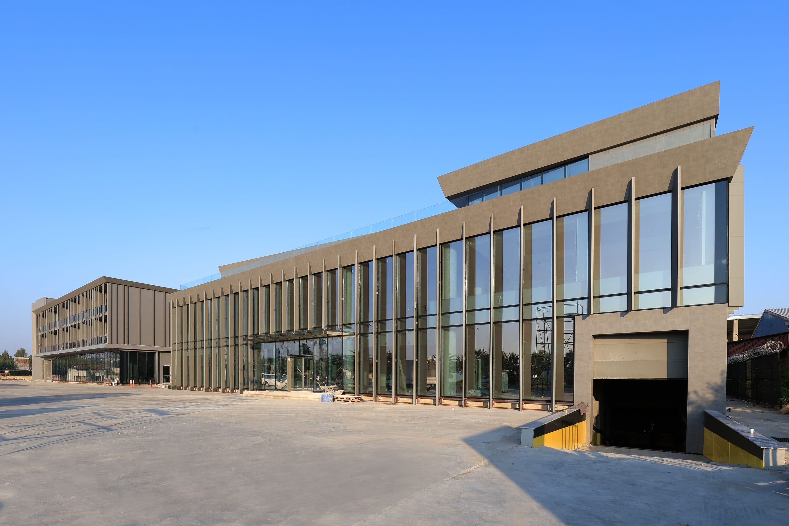 Sanayi tesisinin eğimli saçaklı camlı giriş holü ve arkada ofis bloğu, mavi gökyüzü altında