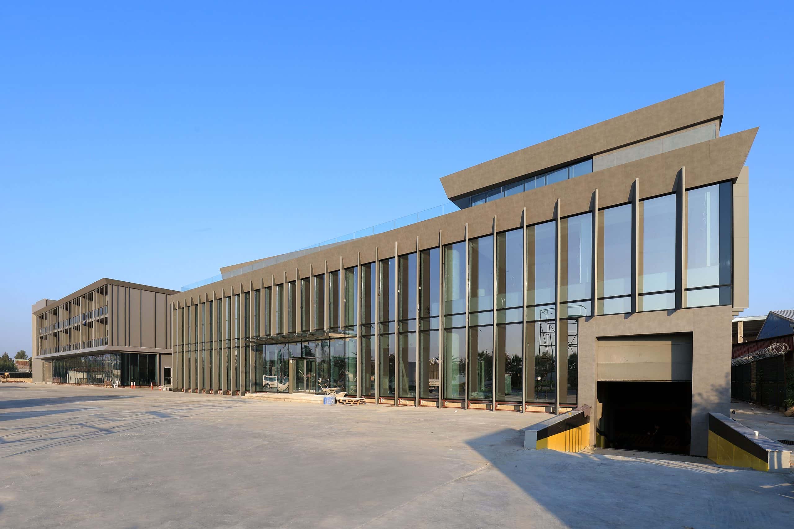 Sanayi tesisinin eğimli saçaklı camlı giriş holü ve arkada ofis bloğu, mavi gökyüzü altında