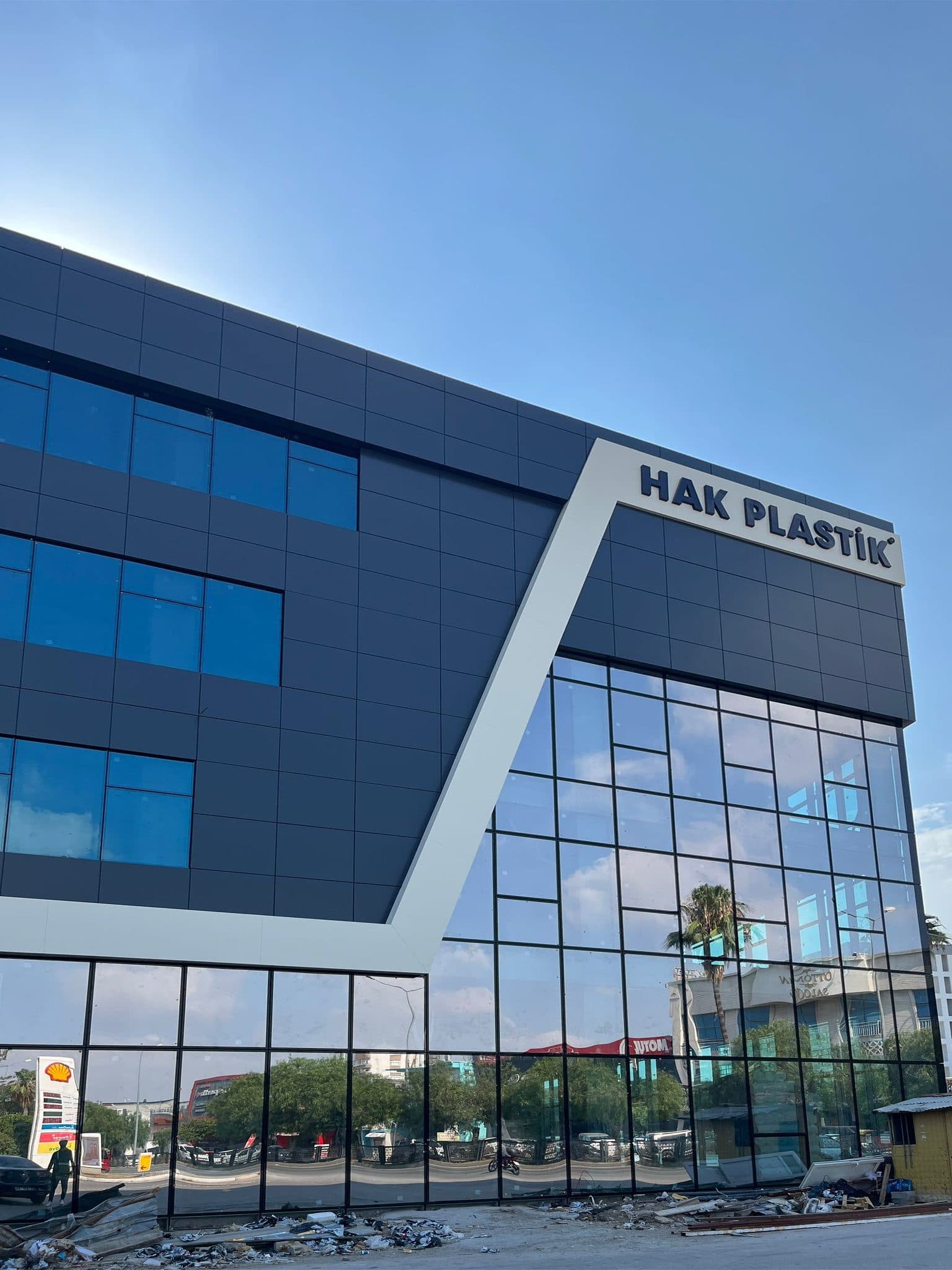 Hak Plastik tabelası ve köşe cephe: beyaz çapraz bant, lacivert kompozit panel, gökyüzünü yansıtan cam