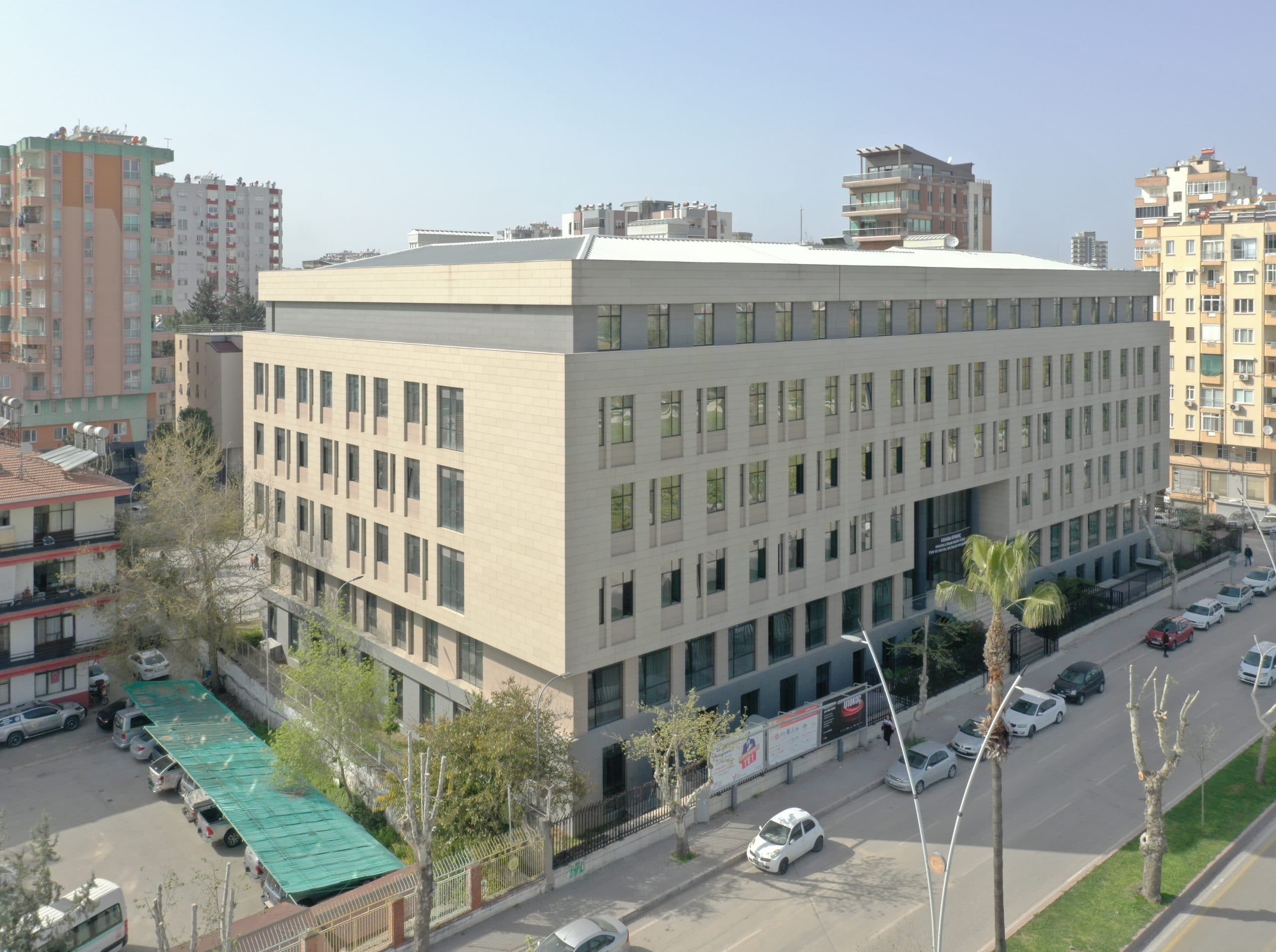 Okulun cadde cephesi ve köşesi havadan; şaşırtmalı derzli açık bej taş görünümlü cephe kaplaması