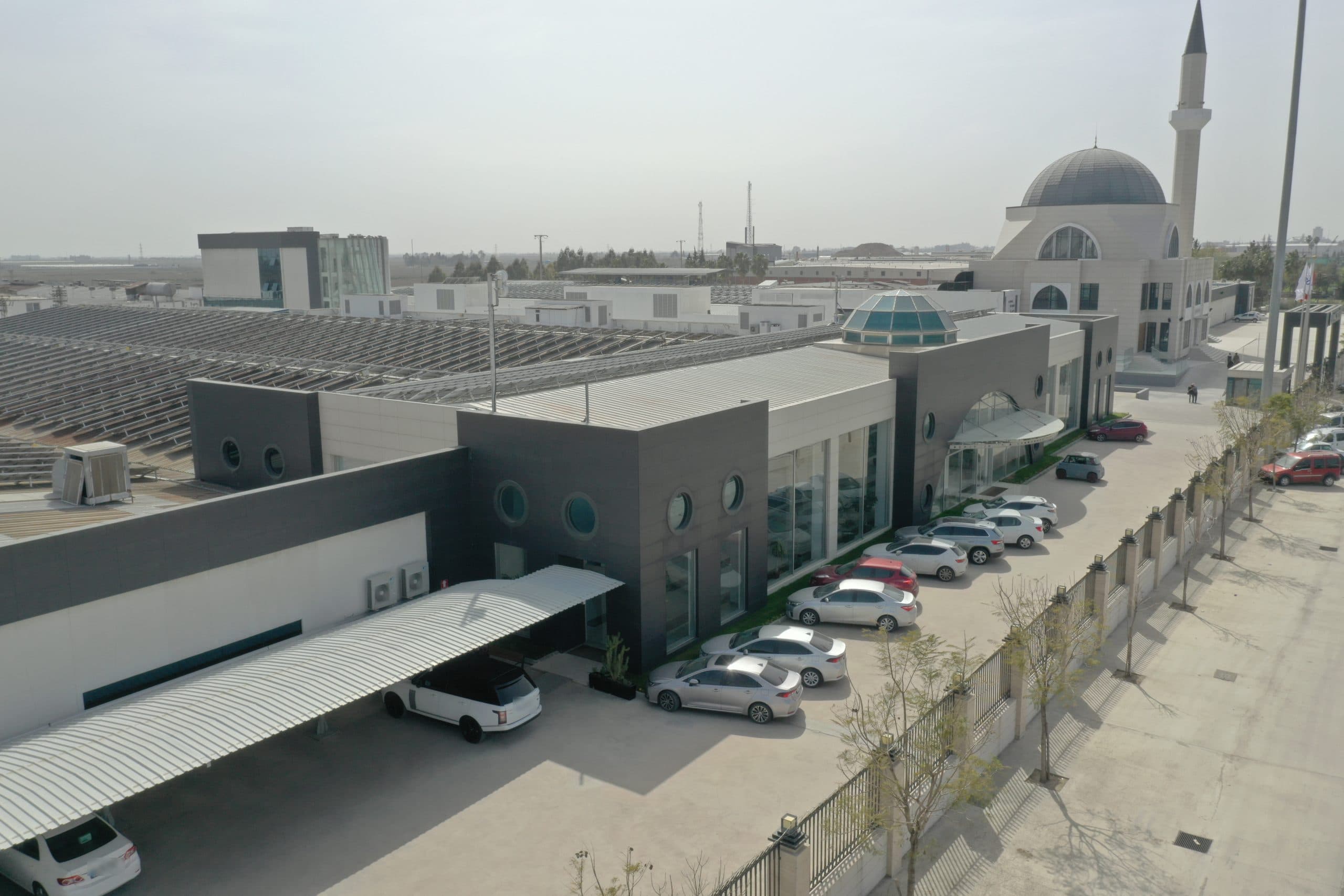 Kıvanç Tekstil idari bloğunun yan cephesi: lombar pencereli antrasit paneller, cam giriş saçağı, arkada cami
