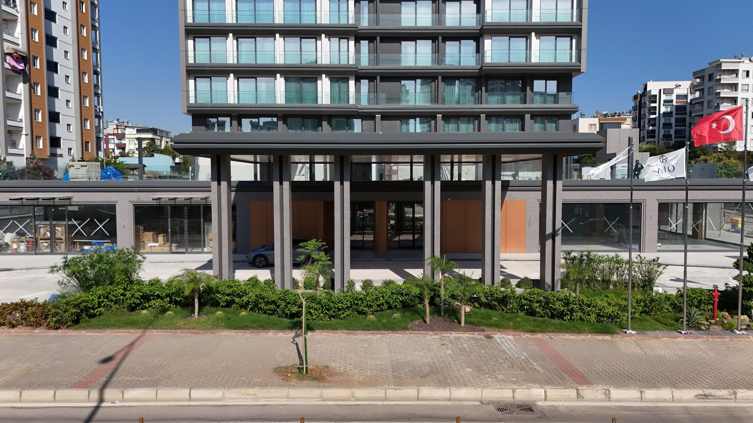 Ramada Plaza giriş saçağı ve podyum katı, cepheden simetrik görünüm, önde peyzaj ve bayraklar