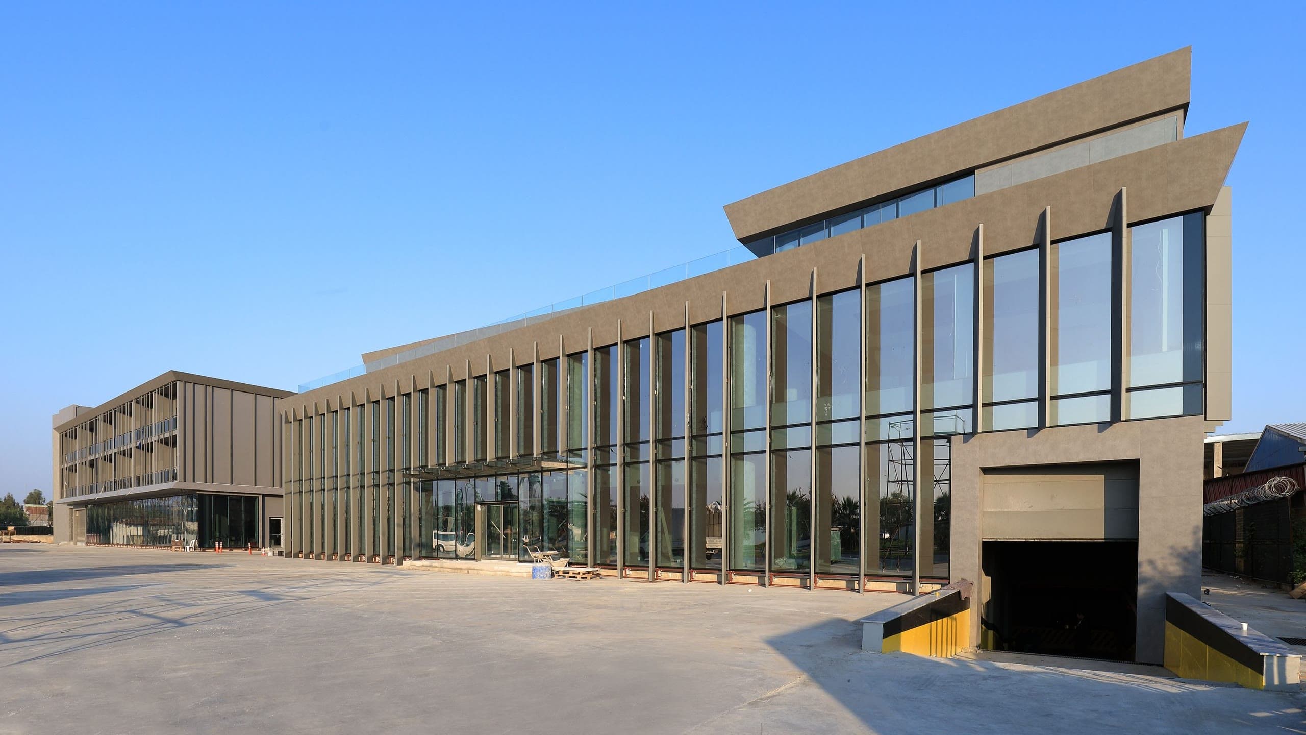 Sanayi tesisinin eğimli saçaklı camlı giriş holü ve arkada ofis bloğu, mavi gökyüzü altında